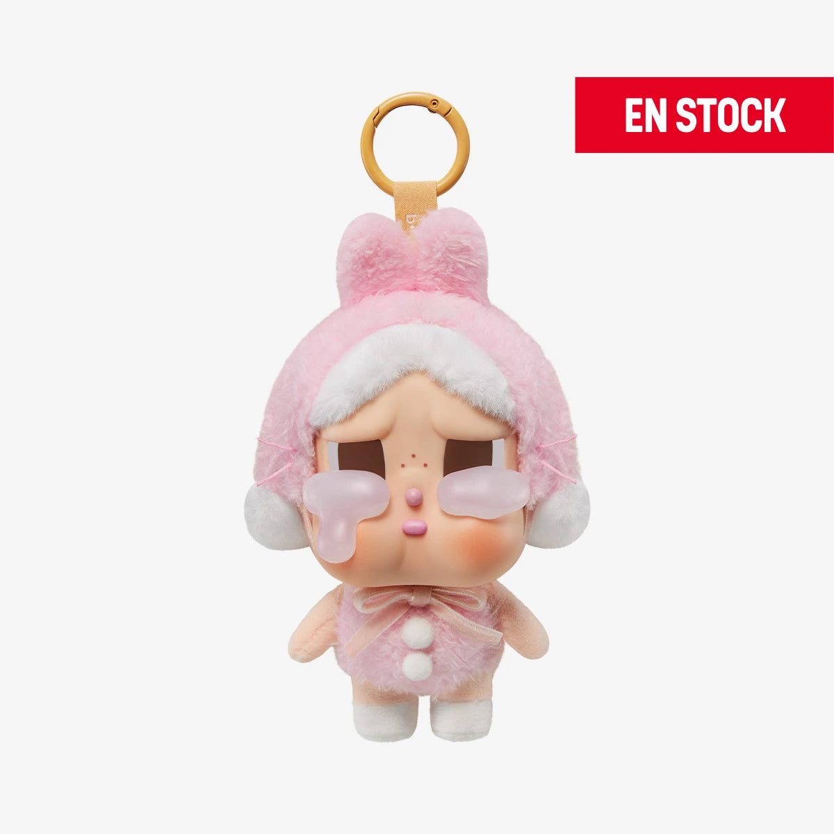 Llavero coleccionable de CRYBABY de la serie Crying Again, con disfraz de peluche rosa con orejas, moño y pompones blancos. El personaje tiene expresión triste con grandes lágrimas transparentes en las mejillas. Cuelga de una anilla dorada con cinta a juego. En la esquina superior derecha aparece una etiqueta roja con el texto EN STOCK