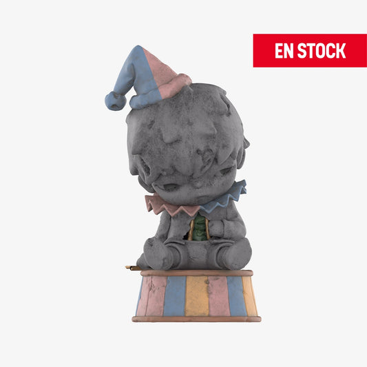 Figura coleccionable de Hirono de la serie Reshape, con apariencia de estatua de piedra deteriorada. Está sentado sobre una base a rayas en tonos pastel azul, rosa y amarillo, con un sombrero de payaso inclinado y cuello de volantes en los mismos colores. La expresión del personaje es melancólica. En la esquina superior derecha aparece una etiqueta roja con el texto "EN STOCK"