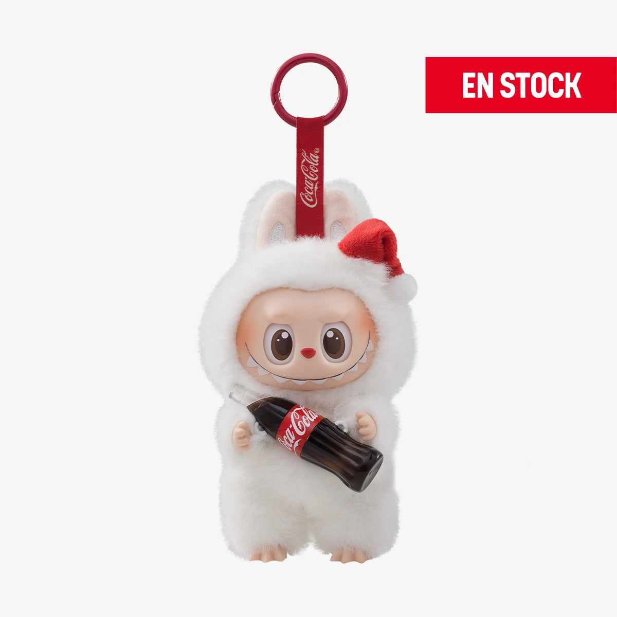 Llavero coleccionable de Labubu en colaboración con Coca-Cola, con traje blanco de peluche con orejas de conejo y gorro navideño rojo. Labubu sostiene una botella de Coca-Cola clásica y cuelga de una anilla roja con cinta de la marca. En la esquina superior derecha aparece una etiqueta roja con el texto "EN STOCK"