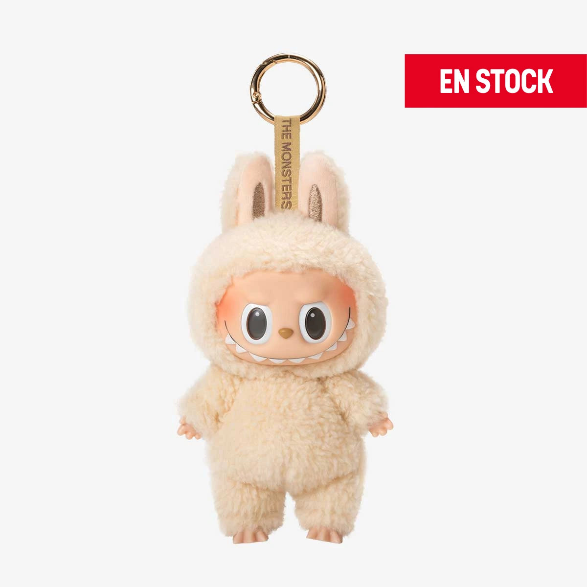 Llavero coleccionable de Labubu de la serie "Macaron", con un adorable traje de peluche color crema que cubre todo su cuerpo, incluyendo orejas de conejo. Tiene una expresión alegre con ojos grandes y brillantes. Cuelga de una anilla dorada con cinta que dice “THE MONSTERS”. En la esquina superior derecha aparece una etiqueta roja con el texto "EN STOCK"