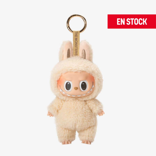 Llavero coleccionable de Labubu de la serie "Macaron", con un adorable traje de peluche color crema que cubre todo su cuerpo, incluyendo orejas de conejo. Tiene una expresión alegre con ojos grandes y brillantes. Cuelga de una anilla dorada con cinta que dice “THE MONSTERS”. En la esquina superior derecha aparece una etiqueta roja con el texto "EN STOCK"