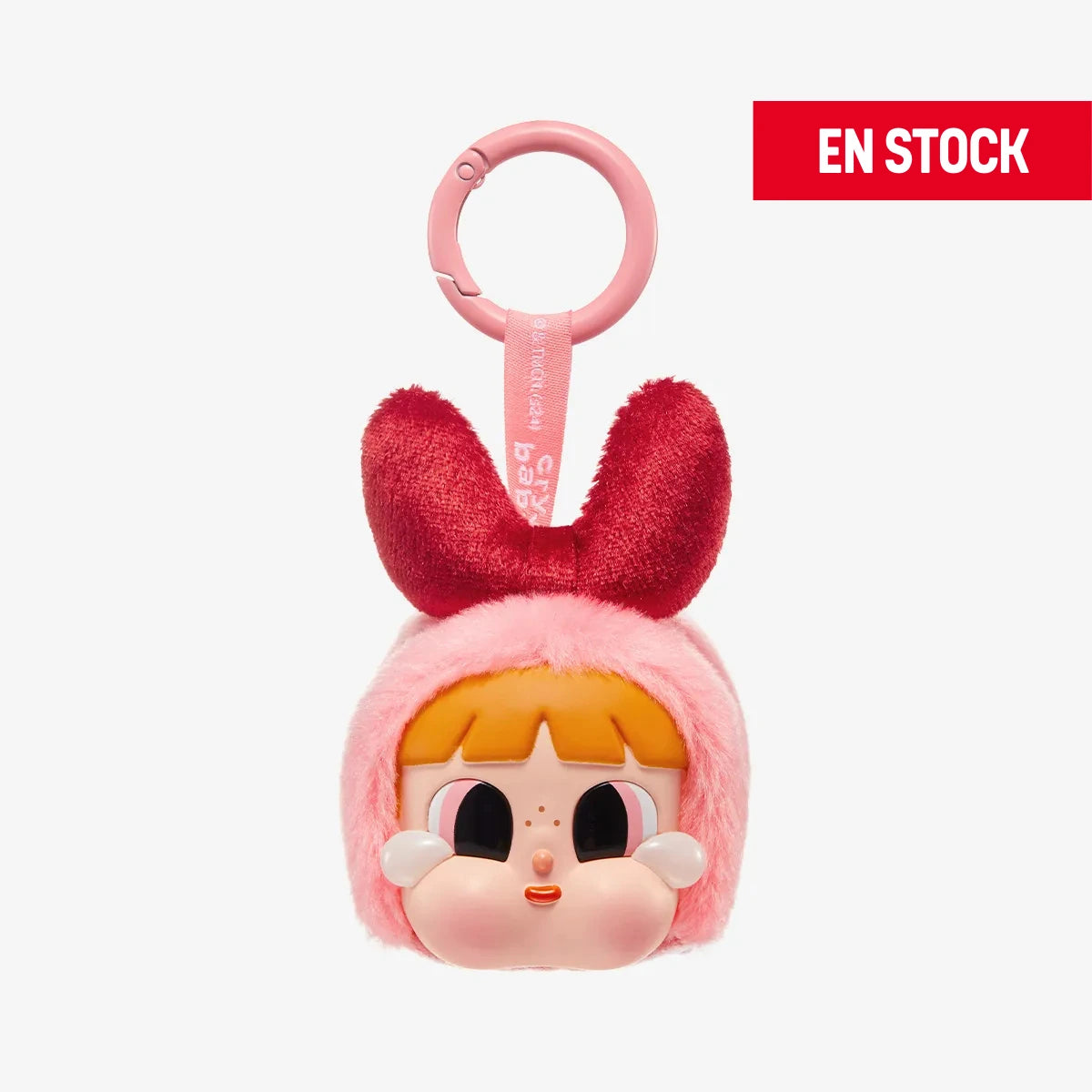Llavero coleccionable de la colaboración CRYBABY × Powerpuff Girls, con cabeza de peluche rosa, lazo rojo de felpa y rostro de personaje con mejillas redondeadas, ojos grandes y cabello naranja. Incluye anilla rosada para colgar y cinta con la marca. En la esquina superior derecha aparece una etiqueta roja con el texto EN STOCK