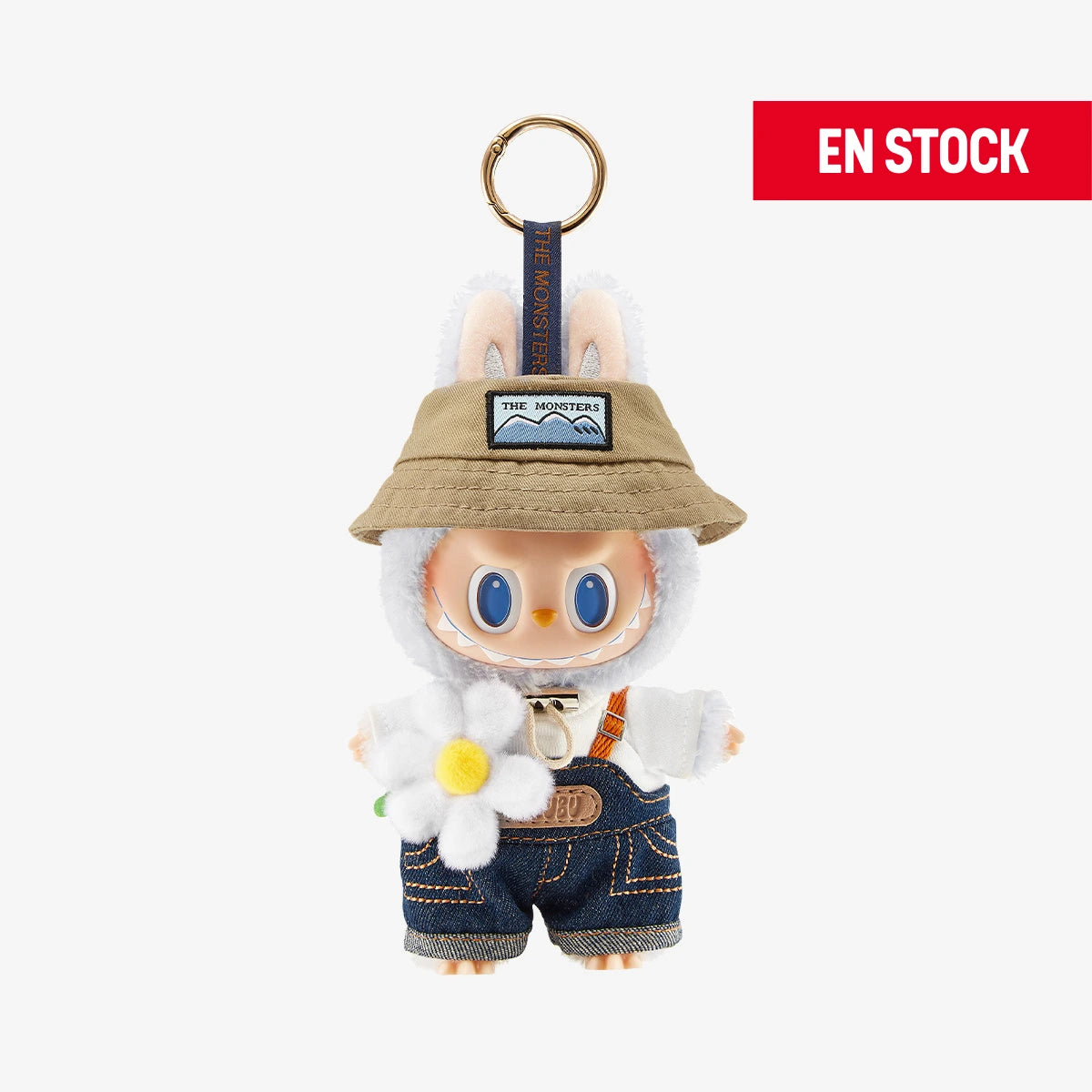 Llavero coleccionable de Labubu de la serie "Fall In Wild", con sombrero beige estilo explorador, camiseta blanca, overol de mezclilla y una flor blanca de peluche en la mano. El personaje tiene grandes ojos azules y orejas de conejo visibles. Cuelga de una anilla dorada con cinta azul que dice “THE MONSTERS”. En la esquina superior derecha aparece una etiqueta roja con el texto "EN STOCK"