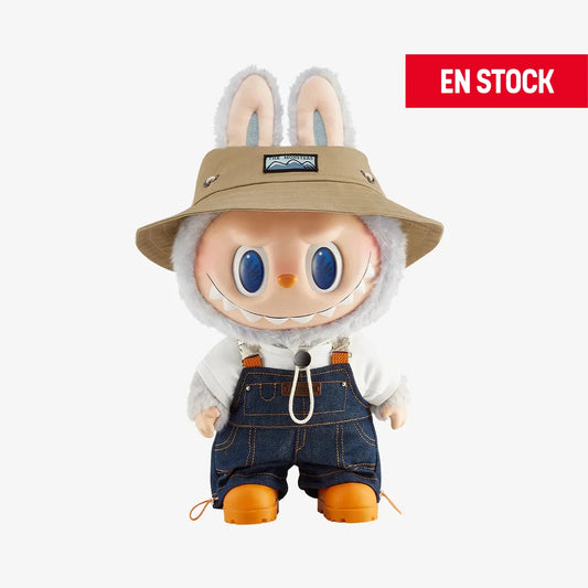 Figura coleccionable gigante de Labubu de la serie "Fall In Wild", con un sombrero beige estilo explorador, overol de mezclilla, camiseta blanca y botas naranjas. El personaje tiene una expresión sonriente con grandes ojos azules y orejas de conejo visibles sobre el sombrero. En la esquina superior derecha aparece una etiqueta roja con el texto "EN STOCK"