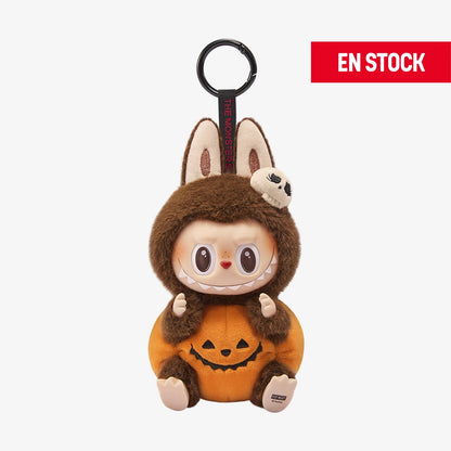 Llavero coleccionable de Labubu de la serie "Happy Halloween", con disfraz de peluche marrón, una calabaza naranja con cara sonriente en el cuerpo y un pequeño cráneo decorativo en la cabeza. El personaje tiene expresión curiosa y grandes ojos marrones. Cuelga de una anilla negra con cinta que dice “THE MONSTERS”. En la esquina superior derecha aparece una etiqueta roja con el texto "EN STOCK"