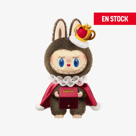 Figura coleccionable de Labubu con temática de San Valentín, vestido como rey con un traje marrón de peluche, capa roja con cuello blanco de volantes y una corona pequeña inclinada sobre la cabeza. Sostiene una caja de terciopelo rojo con la palabra "Labubu" escrita en dorado. En la esquina superior derecha aparece una etiqueta roja con el texto "EN STOCK"