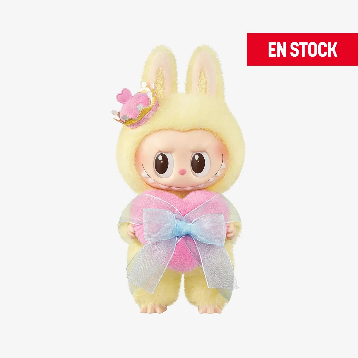 Figura coleccionable de Labubu con temática de San Valentín, usando un traje esponjoso amarillo con orejas de conejo y un pequeño sombrero decorado con corazones. Sostiene un cojín rosa en forma de corazón con un gran lazo azul translúcido. En la esquina superior derecha aparece una etiqueta roja con el texto "EN STOCK"
