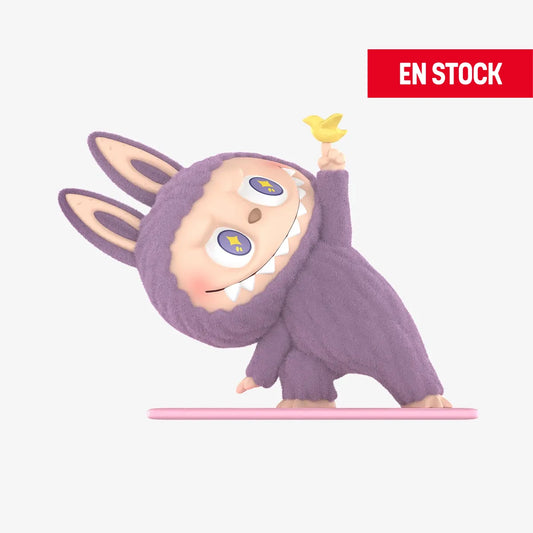 Figura coleccionable de Labubu de la serie Lazy Yoga, en posición de parada de manos con un disfraz morado de conejo peludo y sosteniendo un pequeño pájaro amarillo en un dedo. El personaje tiene ojos con estrellas y una gran sonrisa con dientes afilados. En la esquina superior derecha aparece una etiqueta roja con el texto "EN STOCK".