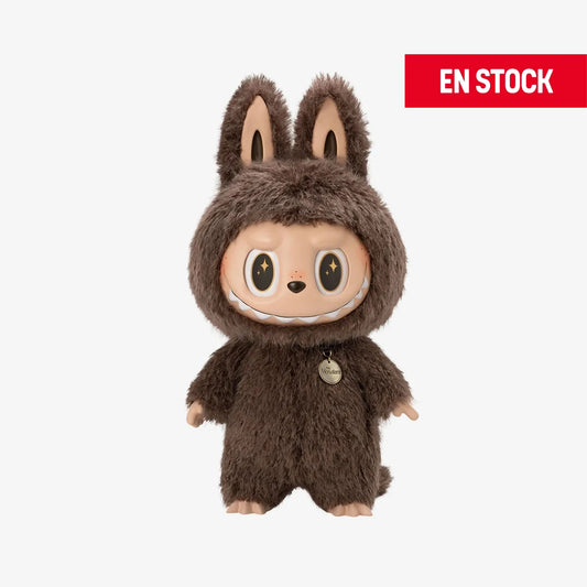 Figura coleccionable de Zimomo con traje completo de peluche marrón oscuro, orejas de conejo y ojos negros con estrellas doradas. Lleva un dije redondo colgando del cuello con la palabra "Wonderland". En la esquina superior derecha aparece una etiqueta roja con el texto "EN STOCK"